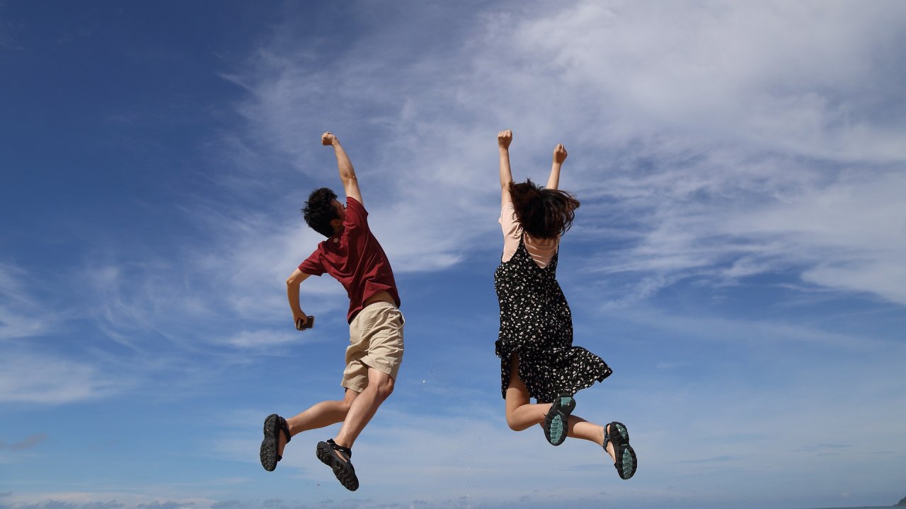 Two students, a male and a female, joyfully jumping in the air against a clear blue sky, celebrating receiving a scholarship.